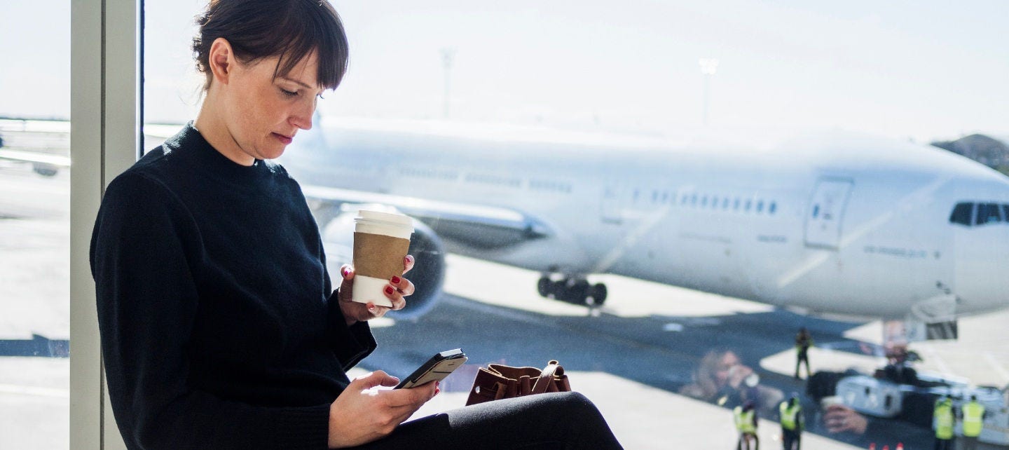 A woman holds a coffee and looks at her phone in front of an airplane. 